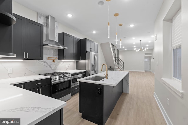 kitchen with a center island with sink, wall chimney range hood, light stone countertops, decorative light fixtures, and stainless steel appliances