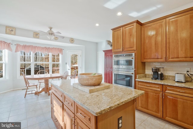 kitchen with decorative backsplash, light stone counters, double oven, ceiling fan, and a center island