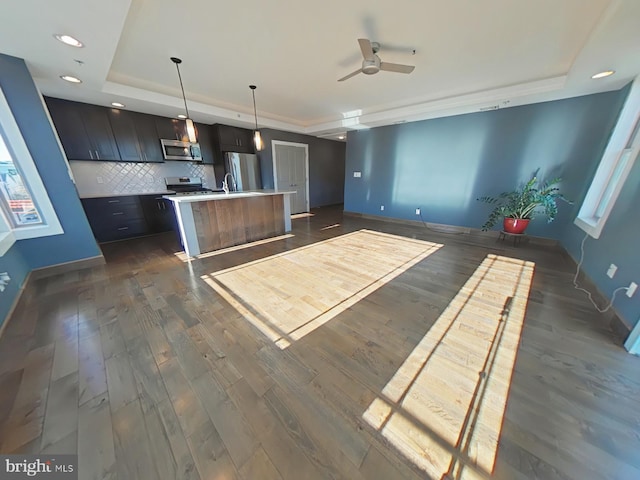 kitchen featuring decorative backsplash, appliances with stainless steel finishes, a tray ceiling, a kitchen island with sink, and pendant lighting