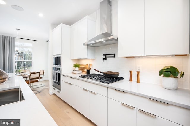 kitchen featuring stainless steel appliances, wall chimney range hood, decorative light fixtures, light hardwood / wood-style floors, and white cabinetry