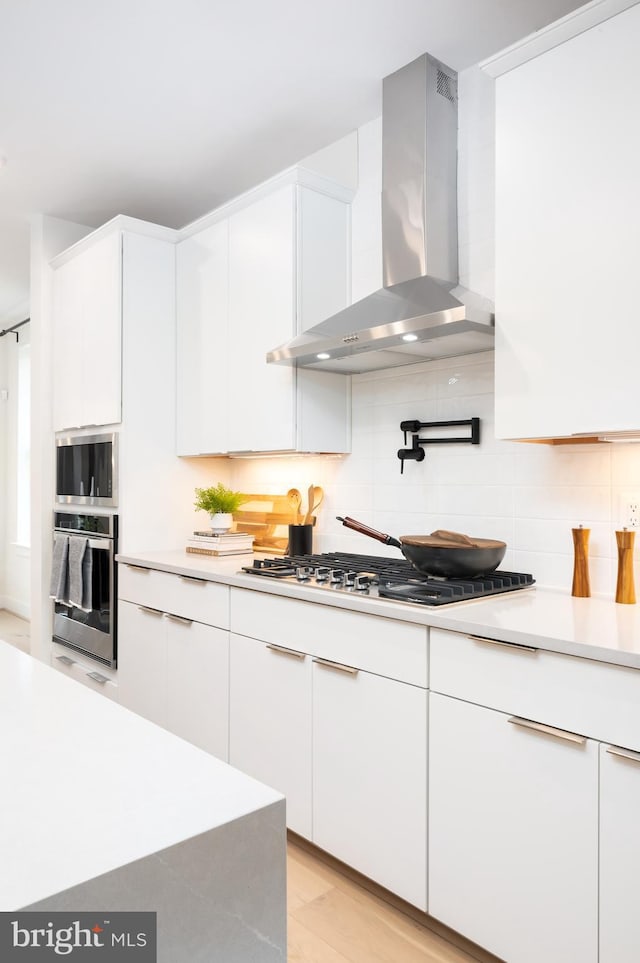 kitchen featuring stainless steel appliances, wall chimney range hood, backsplash, white cabinets, and light wood-type flooring