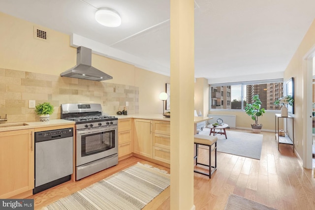 kitchen featuring stainless steel appliances, wall chimney range hood, backsplash, light brown cabinetry, and light wood-type flooring