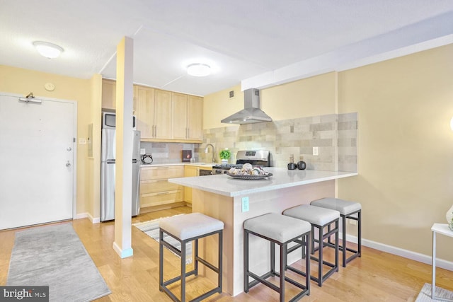 kitchen featuring light brown cabinets, wall chimney range hood, tasteful backsplash, kitchen peninsula, and appliances with stainless steel finishes