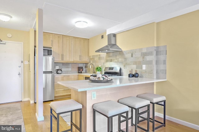 kitchen with a breakfast bar, light brown cabinets, stainless steel appliances, and wall chimney range hood