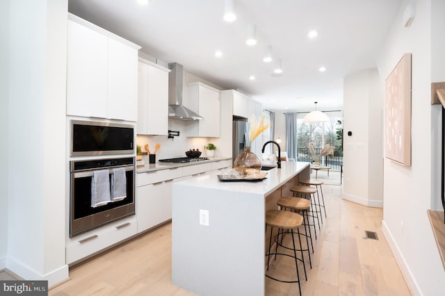 kitchen with stainless steel appliances, white cabinetry, wall chimney exhaust hood, and a center island with sink