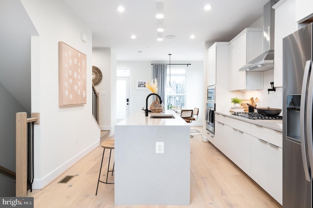 kitchen featuring stainless steel appliances, sink, wall chimney range hood, a center island with sink, and white cabinetry