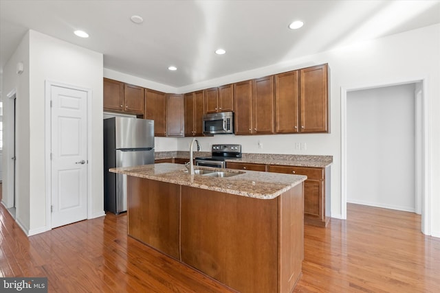 kitchen with stainless steel appliances, light stone counters, light hardwood / wood-style floors, and an island with sink