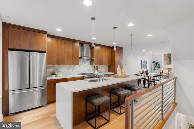 kitchen featuring high end fridge, sink, wall chimney range hood, light hardwood / wood-style floors, and an island with sink