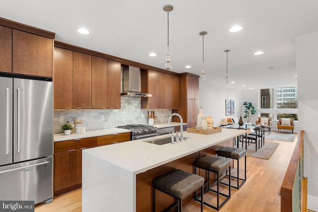 kitchen with wall chimney exhaust hood, high end appliances, a kitchen island with sink, sink, and decorative light fixtures