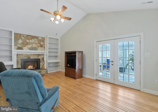 living room with light wood-type flooring, built in shelves, a stone fireplace, french doors, and vaulted ceiling