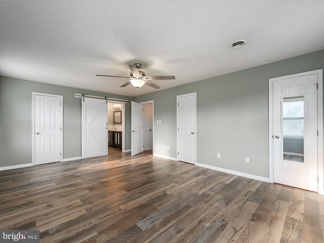 unfurnished bedroom with ceiling fan, dark wood-type flooring, a barn door, connected bathroom, and multiple closets