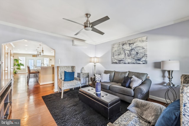 living room with arched walkways, wood finished floors, a ceiling fan, baseboards, and crown molding