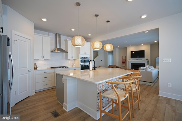 kitchen featuring white cabinets, wall chimney range hood, hanging light fixtures, an island with sink, and stainless steel appliances