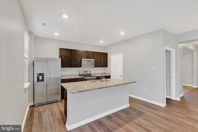 kitchen with a center island with sink, sink, light stone countertops, appliances with stainless steel finishes, and dark brown cabinetry