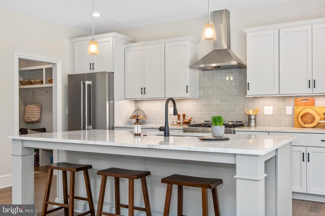 kitchen featuring white cabinets, high end refrigerator, wall chimney exhaust hood, and an island with sink