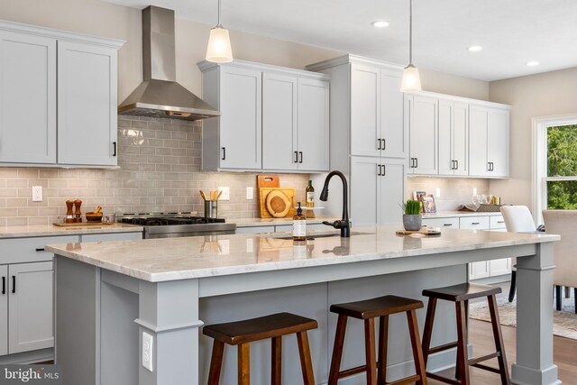 kitchen with stove, a center island with sink, white cabinets, wall chimney exhaust hood, and light stone countertops