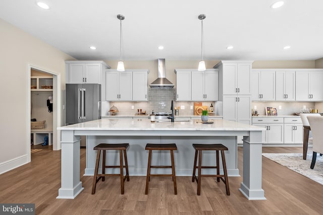 kitchen featuring light wood-type flooring, wall chimney exhaust hood, white cabinets, hanging light fixtures, and an island with sink