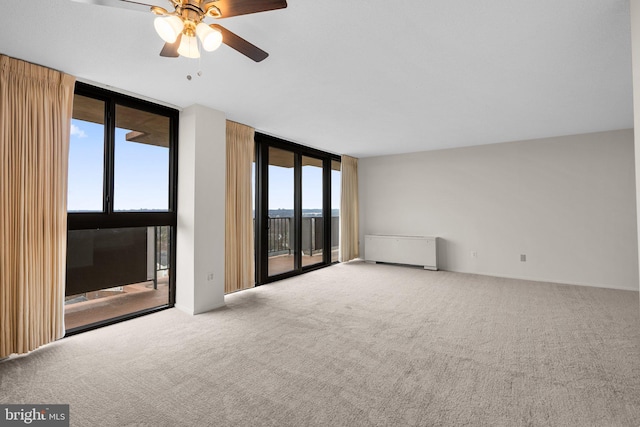 empty room with carpet flooring, ceiling fan, radiator heating unit, and a wall of windows