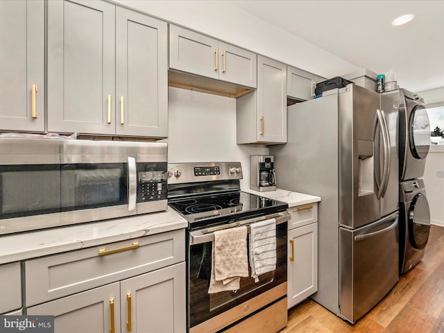 kitchen with appliances with stainless steel finishes, light stone counters, gray cabinetry, light hardwood / wood-style flooring, and stacked washer / dryer