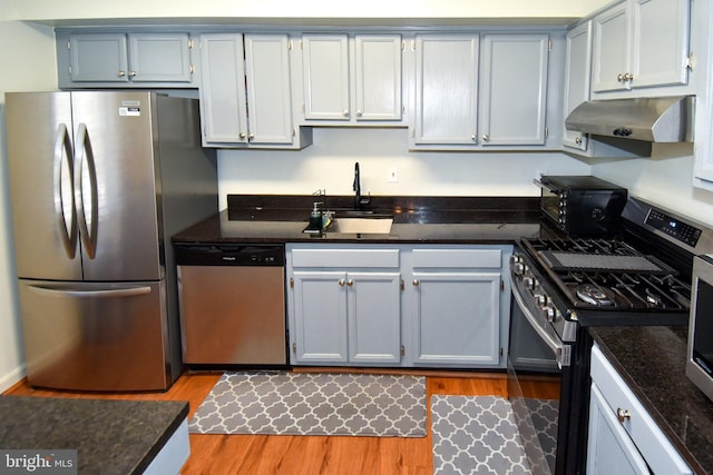 kitchen with appliances with stainless steel finishes, light wood-type flooring, dark stone counters, sink, and exhaust hood