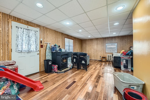 playroom with hardwood / wood-style floors, a wood stove, a drop ceiling, and wood walls