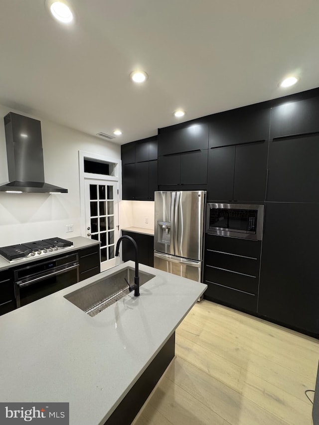 kitchen featuring a center island with sink, wall chimney range hood, sink, light wood-type flooring, and appliances with stainless steel finishes