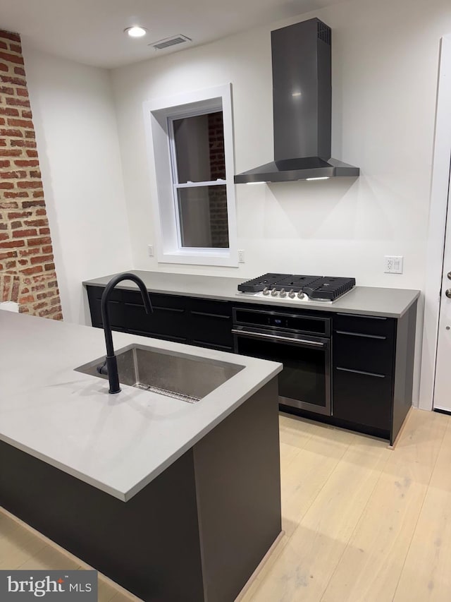 kitchen with kitchen peninsula, appliances with stainless steel finishes, wall chimney exhaust hood, sink, and light hardwood / wood-style flooring