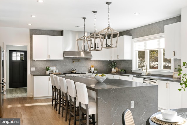 kitchen featuring a center island, white cabinets, wall chimney range hood, sink, and hanging light fixtures