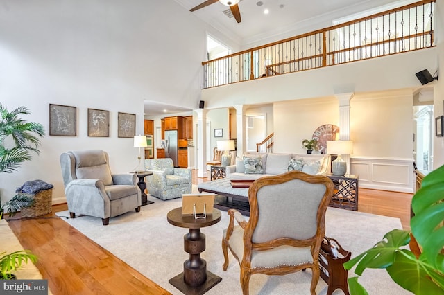 living room featuring a towering ceiling, decorative columns, ornamental molding, ceiling fan, and light hardwood / wood-style floors