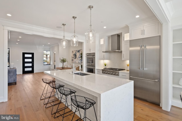 kitchen with decorative light fixtures, stainless steel appliances, a kitchen island with sink, white cabinets, and wall chimney exhaust hood