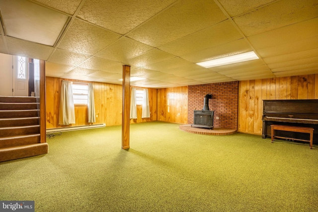 basement featuring carpet flooring, a wood stove, a drop ceiling, a baseboard heating unit, and wood walls