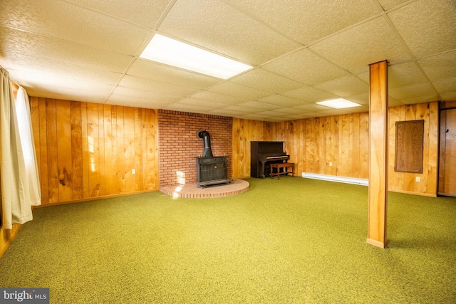 basement featuring a paneled ceiling, wood walls, a wood stove, and carpet floors