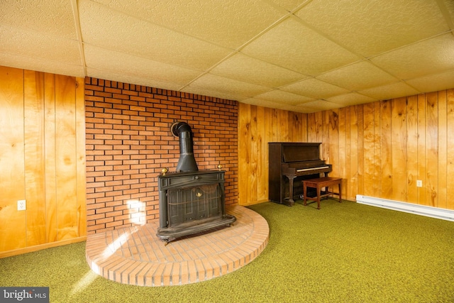 unfurnished living room with carpet flooring, a wood stove, a drop ceiling, a baseboard heating unit, and wood walls