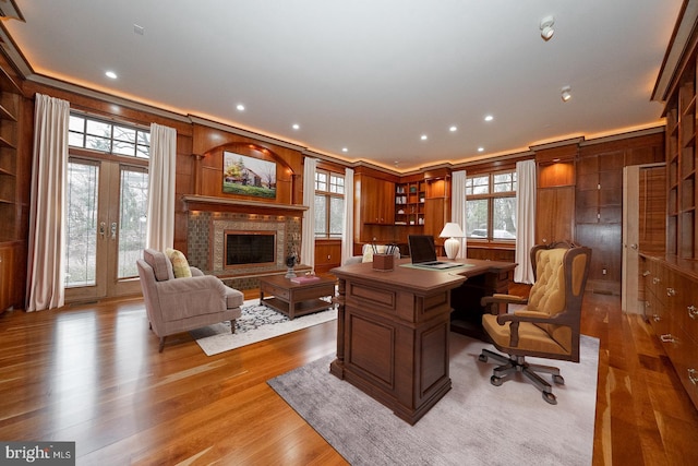 home office featuring a fireplace with raised hearth, wooden walls, french doors, light wood-type flooring, and crown molding