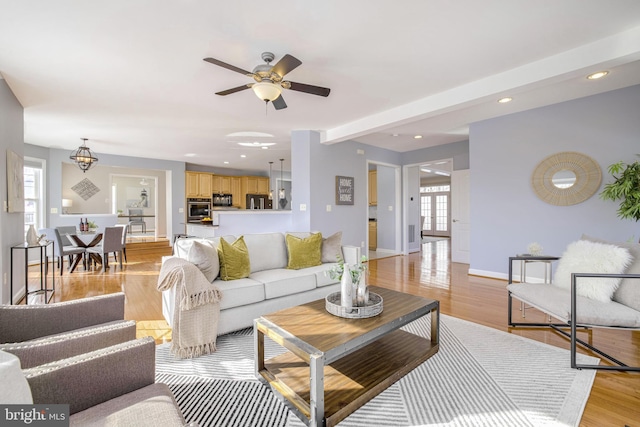 living room featuring beamed ceiling, ceiling fan with notable chandelier, and light wood-type flooring