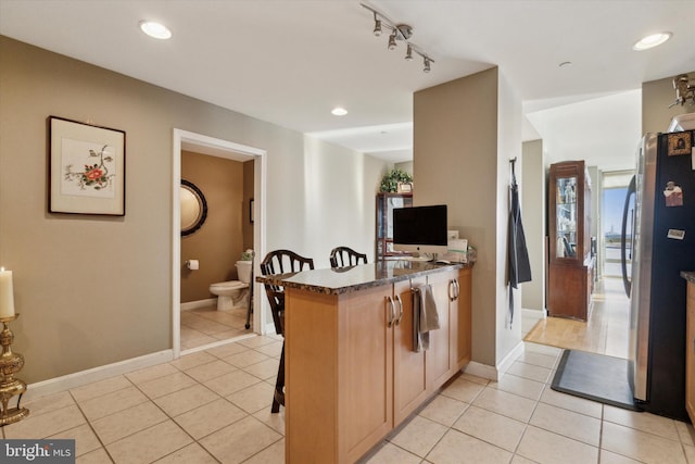 kitchen featuring kitchen peninsula, track lighting, light tile patterned floors, dark stone countertops, and stainless steel refrigerator