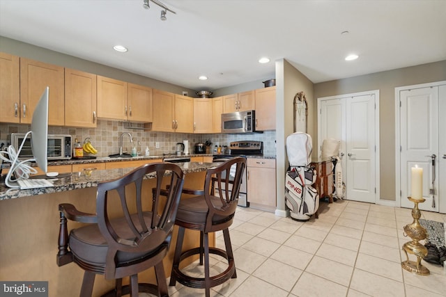 kitchen with light brown cabinetry, dark stone counters, stainless steel appliances, light tile patterned floors, and a breakfast bar area