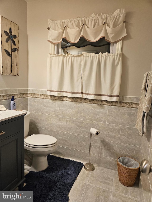 half bath featuring tile patterned floors, a wainscoted wall, toilet, tile walls, and vanity