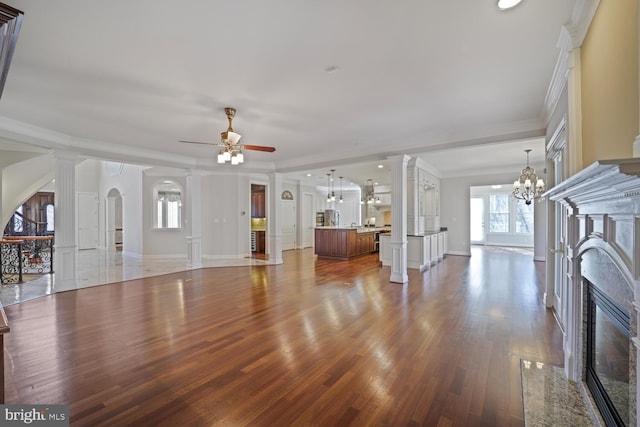 unfurnished living room featuring a premium fireplace, wood finished floors, crown molding, ornate columns, and ceiling fan with notable chandelier