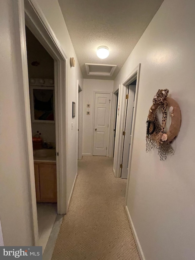 hallway featuring attic access, light carpet, a textured ceiling, and baseboards