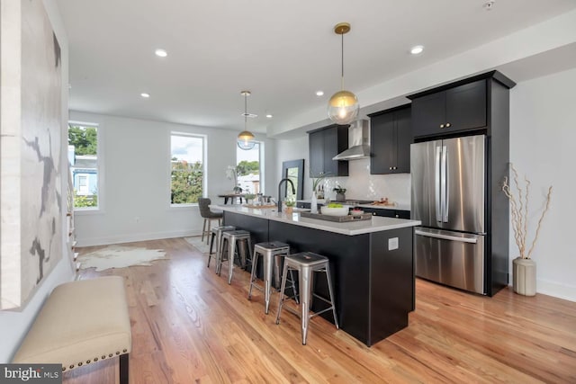 kitchen featuring stainless steel fridge, wall chimney exhaust hood, a breakfast bar, a kitchen island with sink, and hanging light fixtures