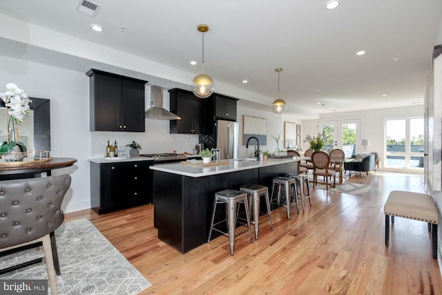 kitchen featuring pendant lighting, a center island with sink, a kitchen breakfast bar, wall chimney exhaust hood, and stainless steel refrigerator