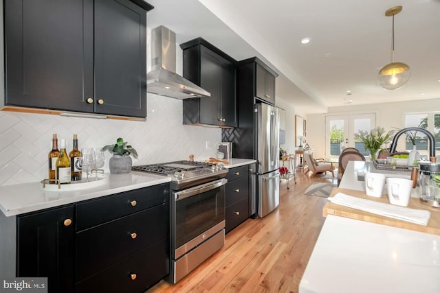 kitchen with wall chimney range hood, hanging light fixtures, light hardwood / wood-style flooring, decorative backsplash, and stainless steel appliances