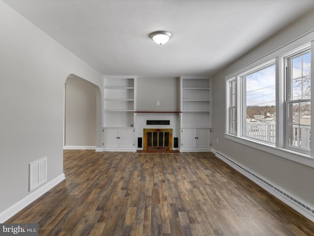 unfurnished living room featuring dark wood-type flooring, built in shelves, a baseboard radiator, and a fireplace