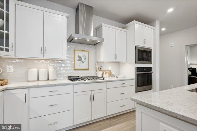 kitchen with light stone countertops, white cabinetry, wall chimney exhaust hood, appliances with stainless steel finishes, and light wood-type flooring