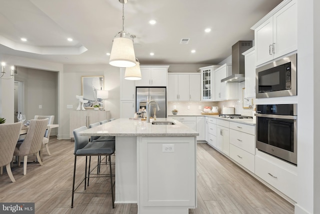 kitchen featuring wall chimney exhaust hood, light hardwood / wood-style flooring, an island with sink, pendant lighting, and appliances with stainless steel finishes