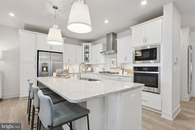 kitchen with sink, stainless steel appliances, wall chimney range hood, an island with sink, and light hardwood / wood-style floors