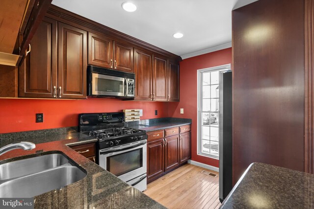 kitchen with appliances with stainless steel finishes, sink, dark stone countertops, ornamental molding, and light wood-type flooring