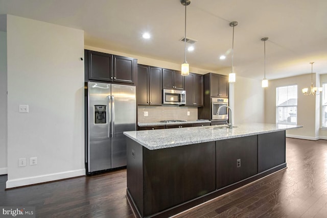 kitchen featuring appliances with stainless steel finishes, an island with sink, hanging light fixtures, light stone counters, and dark brown cabinets