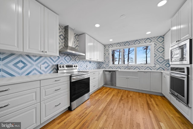 kitchen with backsplash, wall chimney exhaust hood, light wood-type flooring, white cabinetry, and stainless steel appliances